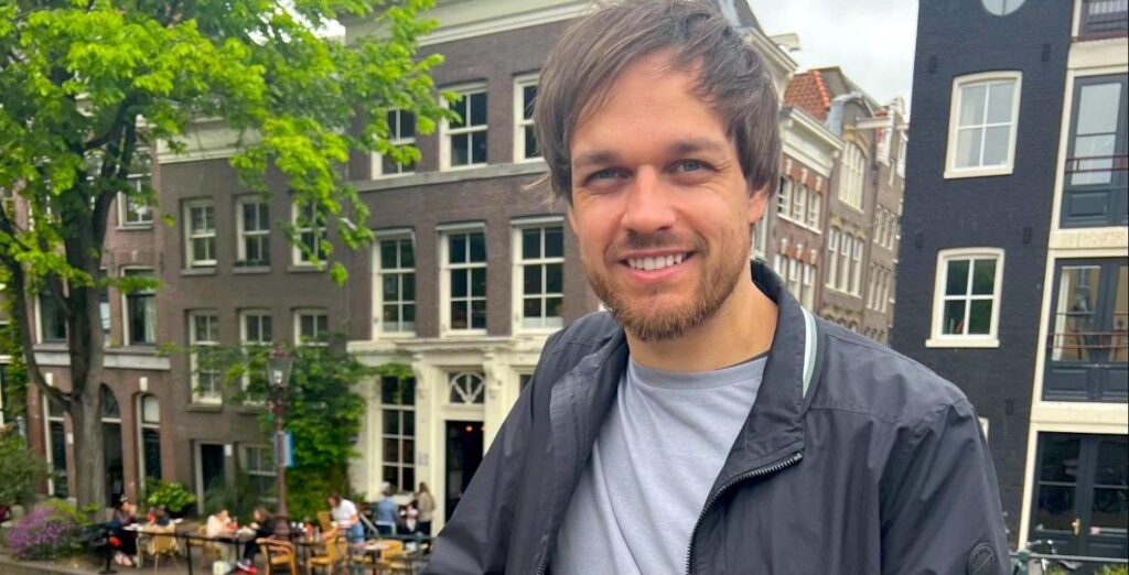 David, a white young man with brown hair, stands on a bridge over an Amsterdam canal.