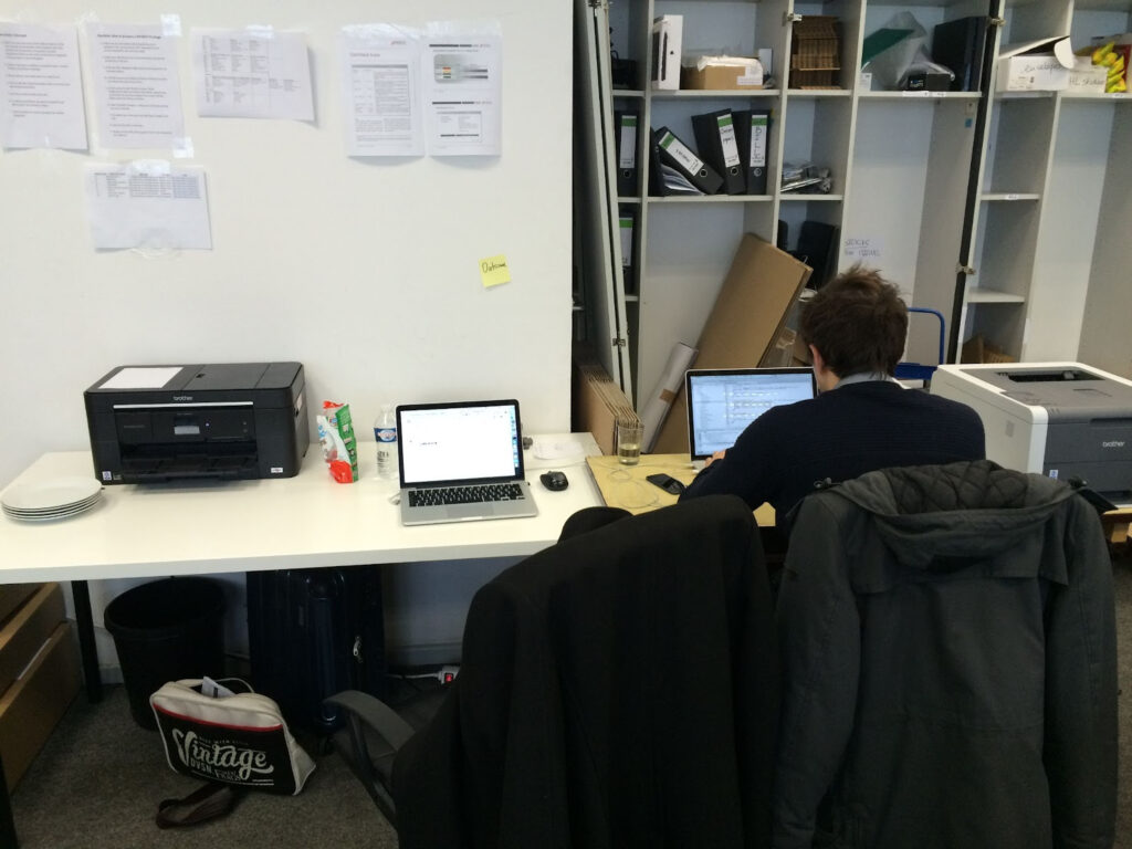 A primehammer engineer sits at a long white desk with his back to the camera, working at a laptop. On the desk, there are another laptop and a printer to his left, and a printer to his right. Behind the desk, there's a white wall with papers and memos stuck to it, and to the right a doorless closet containing folders and boxes.