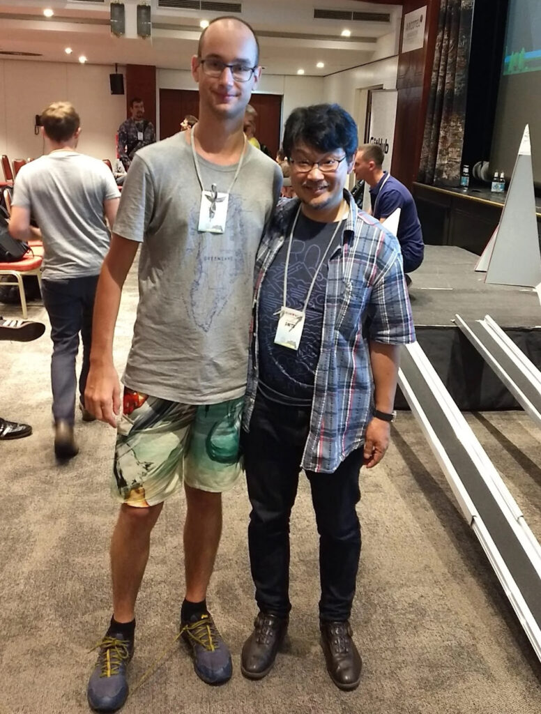 A primehammer engineer (a tall white man with a shaved head and glasses, wearing a grey T-shirt and colorful shorts) poses for a picture next to Matz (a shorter man wearing an open checked shirt on top of a navy T-shirt and dark trousers).