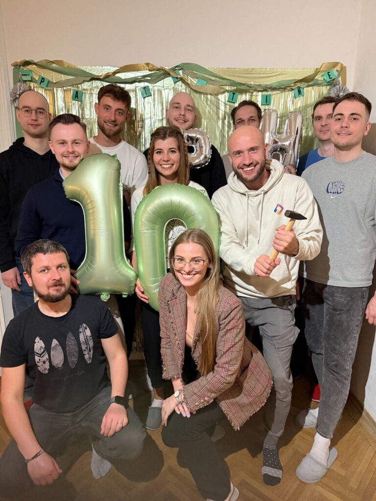 Eleven primehammer people pose for a group photo holding two green ballons (one shaped like a number 1, and the other shaped like the number 0, to make up the number 10).
