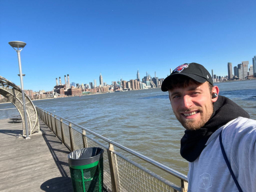 Selfie of Daniel, a young white man wearing a black cap and smiling, posing on a harbor on the river, in front of the New York skyline.